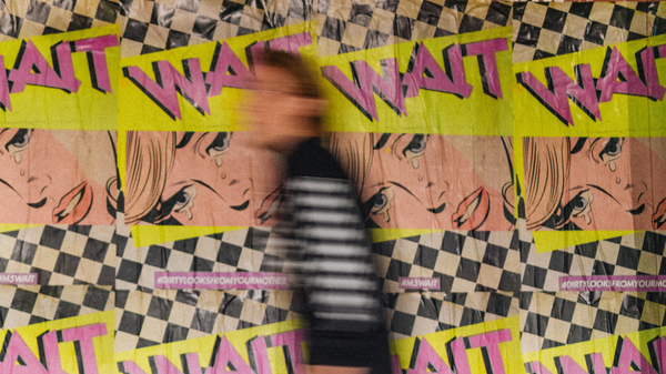 Blurry image of a person walking past a wall papered with posters that read "WAIT"